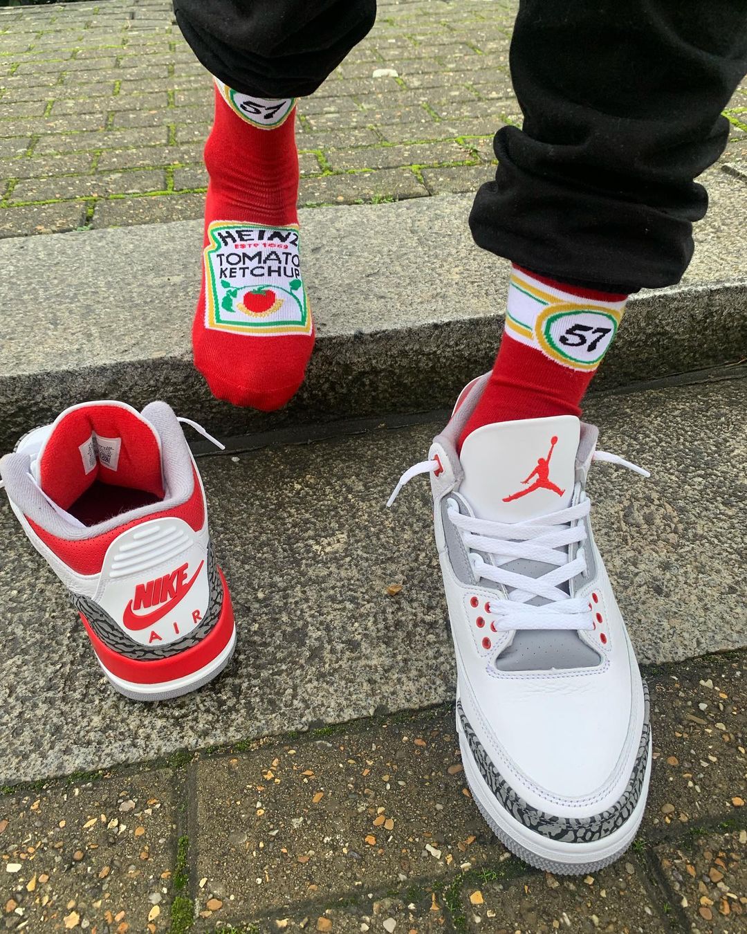 A person wearing red Heinz Tomato Ketchup novelty socks paired with white Air Jordan 3 Fire Red sneakers while standing on concrete steps
