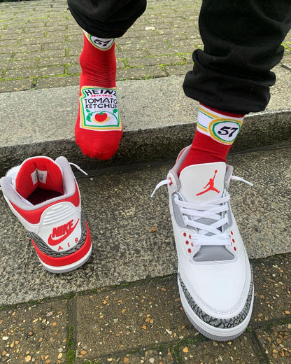 A person wearing red Heinz Tomato Ketchup novelty socks paired with white Air Jordan 3 Fire Red sneakers while standing on concrete steps